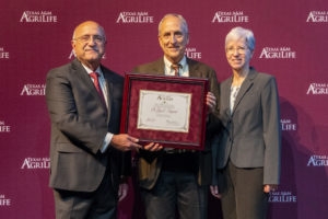 man with white hair and suit accepting award from man in gray suite and glasses and woman in gray suit and glasses