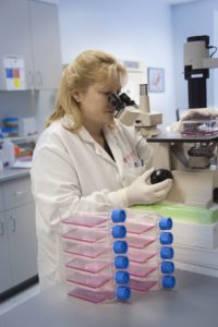 Blonde woman in lab coat looks at diagnostic media under a microscope