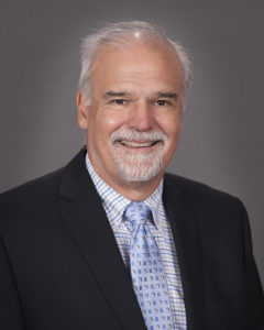 Portrait of a man with grey hair and mustached in a black suit with blue, white, and tan patterned button down and blue and silver square designed tie.