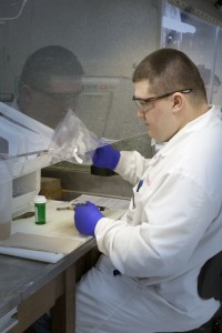 Man in lab coat and goggles working with sample from a plastic Ziploc in biosafety cabinet