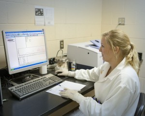 A blonde haired woman in lab coat and gloves analyzing data on computer