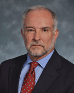 A portrait of a grey haired man in a black blazer, blue button down, and red tie.