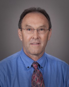 Portrait of a man with dark hair and glasses in a blue button down and maroon and grey designed tie