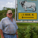 Man in a blue, tan and maroon patterned button down, cap, and glasses smiling as he stands in front of a blue and yellow J.D. Hudgins, Inc. Brahmans sign surrounded by trees and plants.