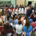 Large group of students in white aprons examining contents on a metal tray in a lab with an instructor
