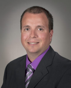 Portrait of man with short hair in a black suit with a purple button down and grey and purple striped tie