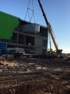 Construction work being done for a new building surrounded by mud. Large yellow crane is installing three large air units into the building structure.