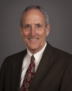 Portrait of man with short grey hair with a brown suit, white button down and maroon, tan, and navy designed tie.