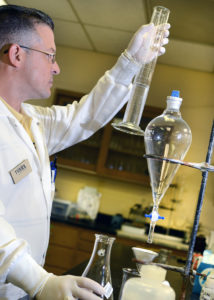 Man in a white lab coat and gloves examines the amount of fluid in a large glass graduated cylinder at a lab bench with other glass equipment.