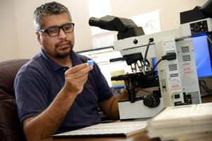 man with gray hair and black glasses evaluating glass side beside a microscope