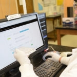 Man with sample plate in white gloves and lab coat sitting at computer