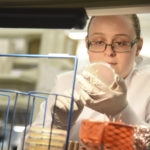 Woman with brown hair in pony tail and glasses examining a Petri dish while wearing white gloves and lab coat