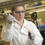 Woman with brown hair, white gloves, white gown, and blue safety glasses pipetting brown liquid in tube