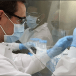 Man with brown hair wearing mask, white gown, and blue gloves pipetting liquid into tube