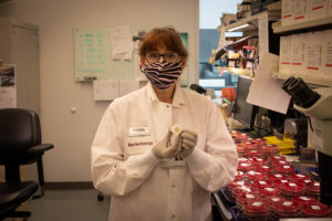 Woman with red hair and black glasses wearing a pink and black zebra print face mask. She is wearing a white lab coat and holding a small circular pin.