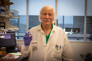 Man with white hair and a white lab color holding a small circular pin with purple gloves.