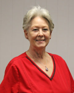 Woman with gray hair and wearing a red scrub top smiling for a portrait in front of a white wall.