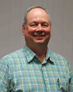 Man with gray hair and a plaid, green, orange, blue, and white shirt smiling for a portrait in front of a white wall.
