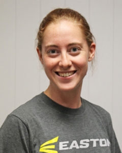 Woman with red hair and a gray t shirt smiling for a portrait in front of a white wall.