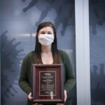 Woman with brown hair, gray face mask, and green sweater poses with an award plaque
