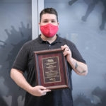 Man with brown hair, red face mask and gray shirt poses with an award plaque