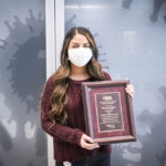 Woman with long curled hair, maroon sweater, and white face mask posing with an awards plaque