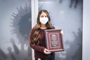 Woman with long curled hair, maroon sweater, and white face mask posing with an awards plaque
