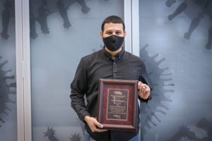 Man with black hair, black face mask, and a black shirt poses with an awards plaque