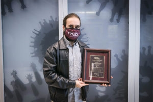 Man with long brown hair, maroon face mask and black leather jacket poses with an awards plaque