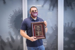 Man with dark hair, maroon face mask, and blue shirt poses with an awards plaque