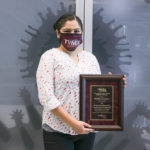 Woman with black hair tied back, maroon mask, and white shirt with pastel colored hearts holds an awards plaque