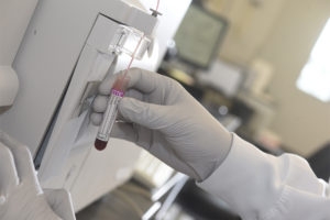 Hand in white glove holding a red blood tube under a machine.