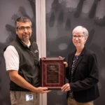 Man wearing a white shirt and black vest accepting plaque from woman with white hair and black suit