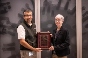 Man wearing a white shirt and black vest accepting plaque from woman with white hair and black suit