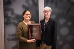 Woman with brown hair and a green sweater accepting a plaque from a woman with white hair and a black suit