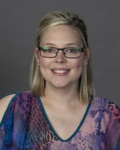 woman with blonde hair and black glasses posing for a professional portrait while wearing a floral blue and pink shirt
