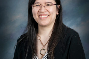 woman with black hair, glasses, and a black blazer coat smiling for a professional portrait