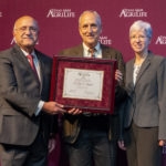man with white hair and suit accepting award from man in gray suite and glasses and woman in gray suit and glasses