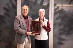 man with white hair and gray suit jacket accepting award from woman in white vest and maroon shirt