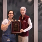 woman white white shirt and overalls accepting award from woman in maroon vest and white hair