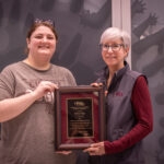 woman in maroon shirt and gray vest presenting award plaque to woman in gray shirt and brown hair