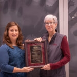 woman in maroon shirt and gray vest presenting award plaque to woman in blue shirt and brown hair