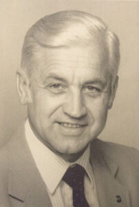 black and white professional portrait of man with white hair and black tie and gray coat.
