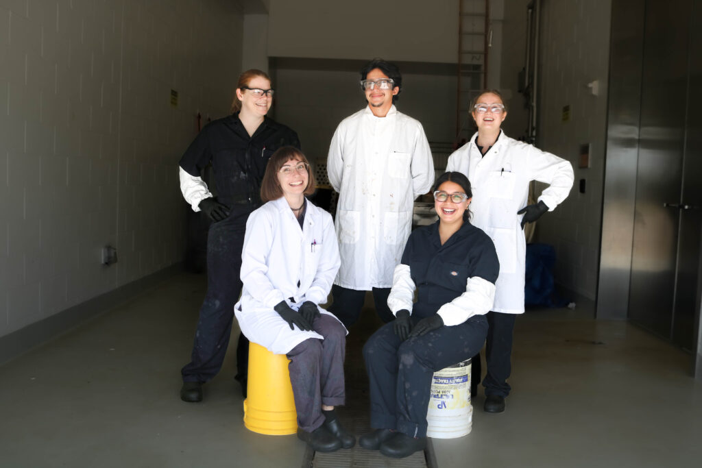 Five people in lab coats and coveralls pose for a group photo