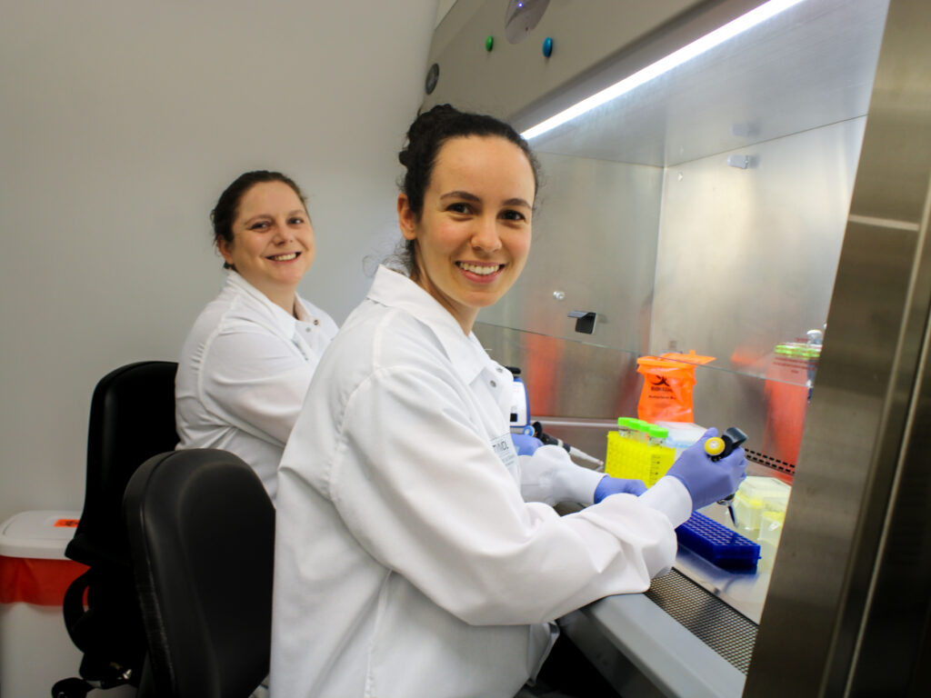 two women in white lab coats sit by a lab hood and smile for a portrait