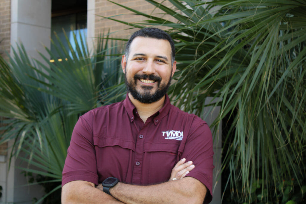 man in maroon shirt and black hair and beard standing in front of palm tree with arms crossed